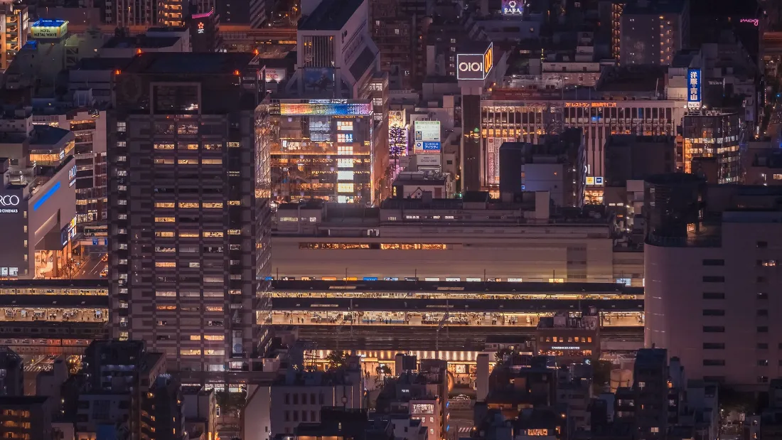 高空俯瞰东京城市夜景，密集楼宇灯火通明，横向高架轨道列车穿行其间。