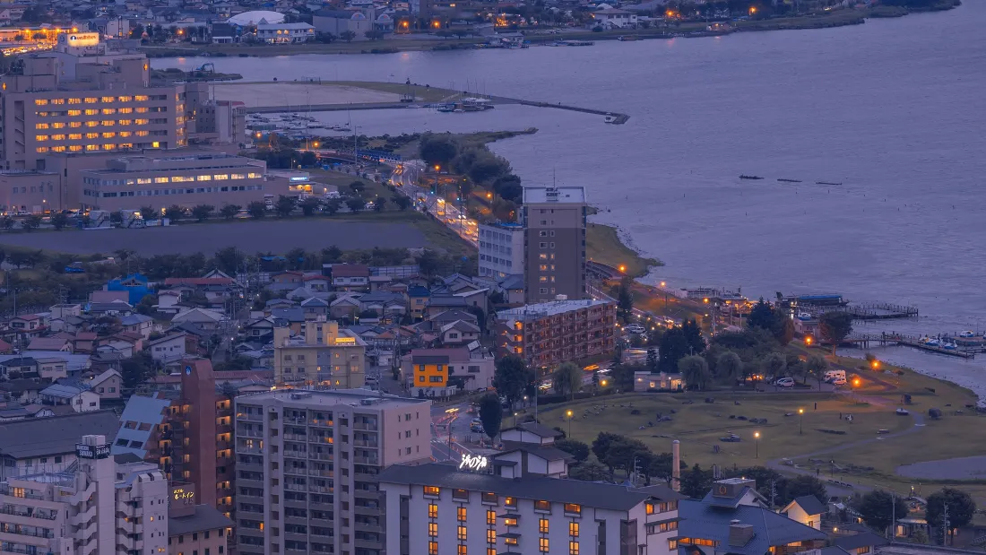 2k城市夜景壁纸|都市风景背景图片 - 建筑水域「哲风壁纸」 黄昏时分的滨水城市俯瞰景观,沿岸建筑亮起暖黄灯光,水面泛着淡紫暮色。