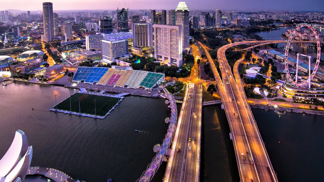 航拍黄昏入夜的城市滨水夜景，高架道路车流灯光蜿蜒，摩天轮与摩天楼林立。