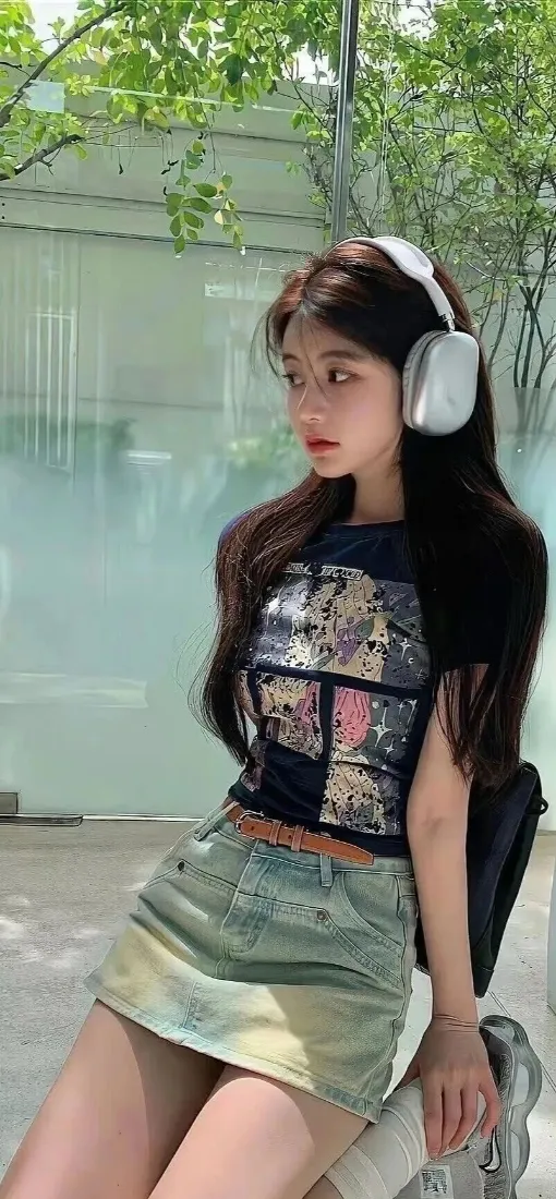 户外绿植玻璃墙前，戴白色头戴耳机的长发女生穿印花上衣与牛仔短裙侧身倚靠