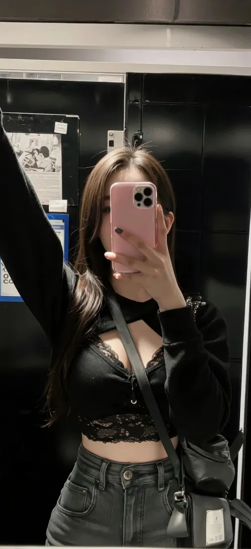 长发女子在镜前用粉色手机自拍，身着黑色露腰上衣与牛仔裤。