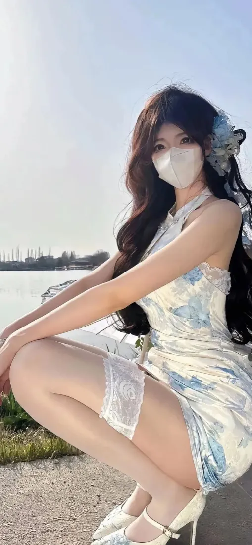 戴口罩长发女子着蓝白挂脖旗袍，蹲在水边户外，背景是蓝天与远岸。