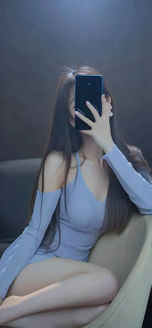 长发女子穿浅蓝色露肩紧身衣坐沙发上，举手机遮挡面部自拍