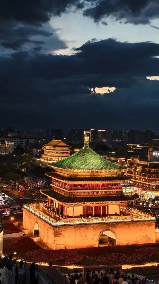 黄昏时分，西安钟楼被暖黄灯光点亮，周边城市灯火璀璨，天空云层厚重。