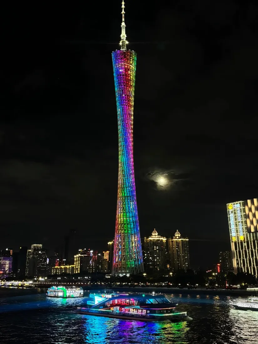 夜晚珠江畔，彩虹灯光点亮的广州塔矗立，江面游船倒映着霓虹光影。