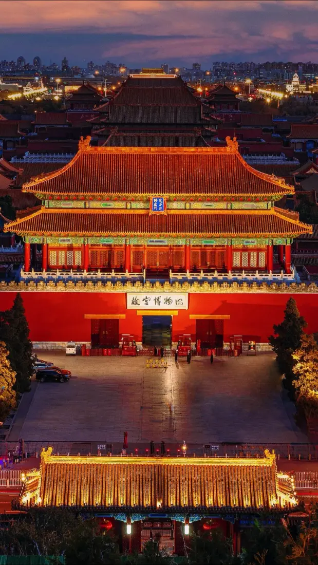 黄昏时分的故宫神武门，红墙金瓦被暖光点亮，远处城市灯火与晚霞相映。