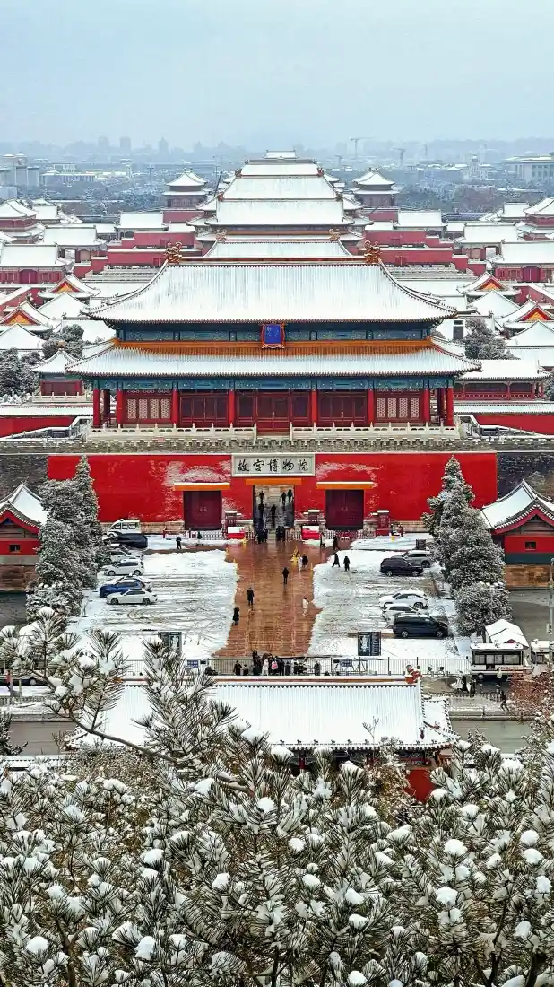 雪后俯瞰故宫，红墙金瓦覆满白雪，中轴建筑对称延伸，游人漫步广场。