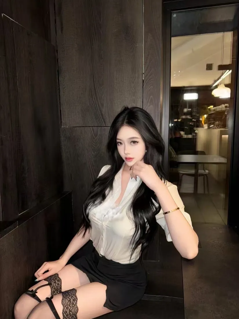 长发白衣黑裙黑丝女性坐于深色室内，手托脸颊看向镜头。