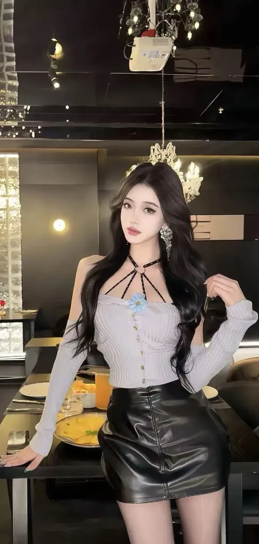 长发美女身着灰色露肩上衣与黑色皮裙，站在灯光奢华的室内酒吧餐桌旁。