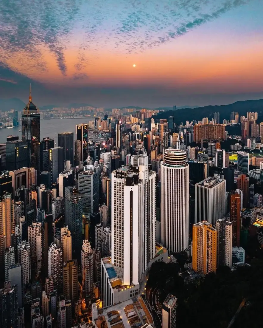 黄昏时分高空俯瞰的密集都市摩天建筑群，远处可见水域与山峦，天空晕染橙蓝渐变晚霞