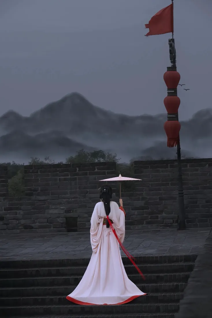 烟雨朦胧的古城墙石阶上，白衣古装女子撑油纸伞背对镜头望向雾中远山。