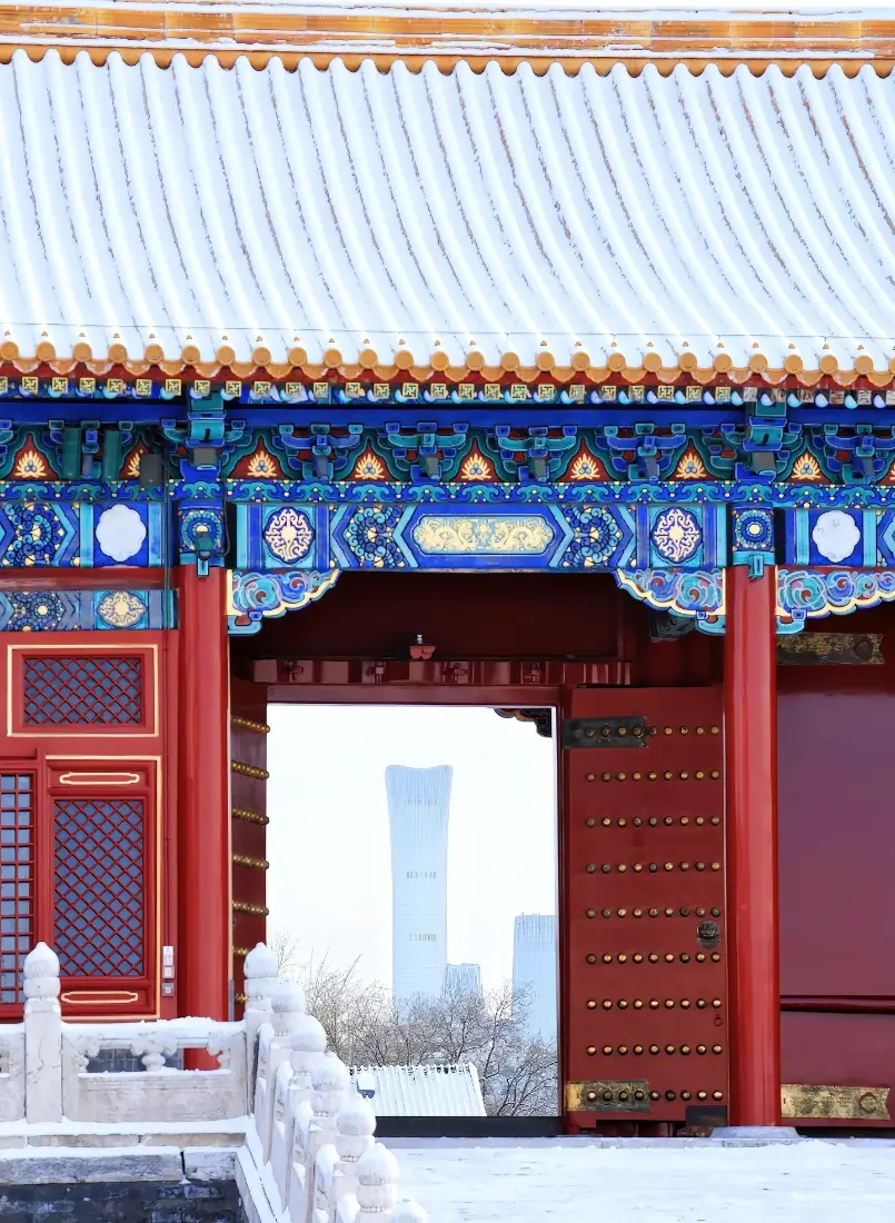 雪后故宫宫门，红墙彩瓦覆雪，门洞框出远处现代摩天楼。
