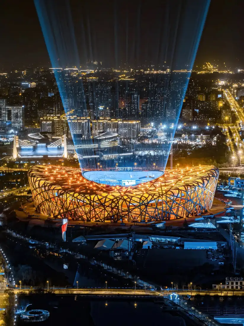 航拍视角下夜晚的北京鸟巢，金色灯光照亮钢结构，上方投射蓝色光束，背景是城市夜景。
