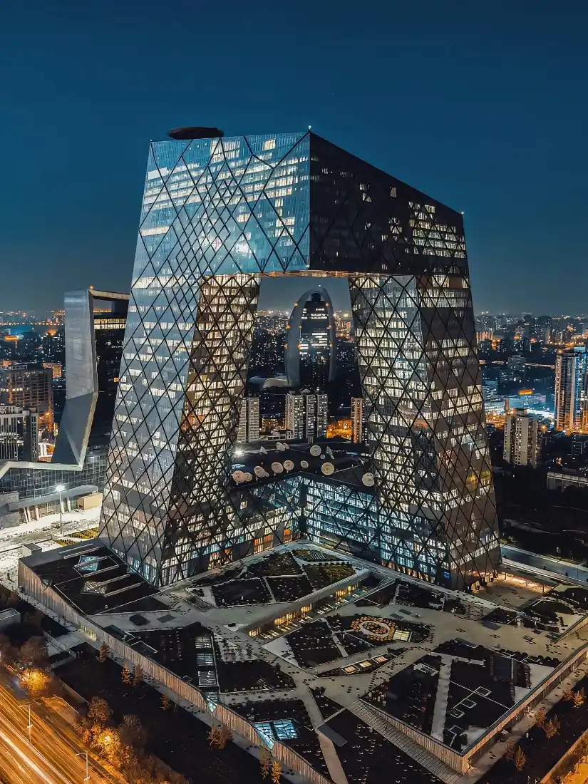北京央视大楼夜景，玻璃幕墙灯光璀璨，周边城市建筑群灯火点缀。