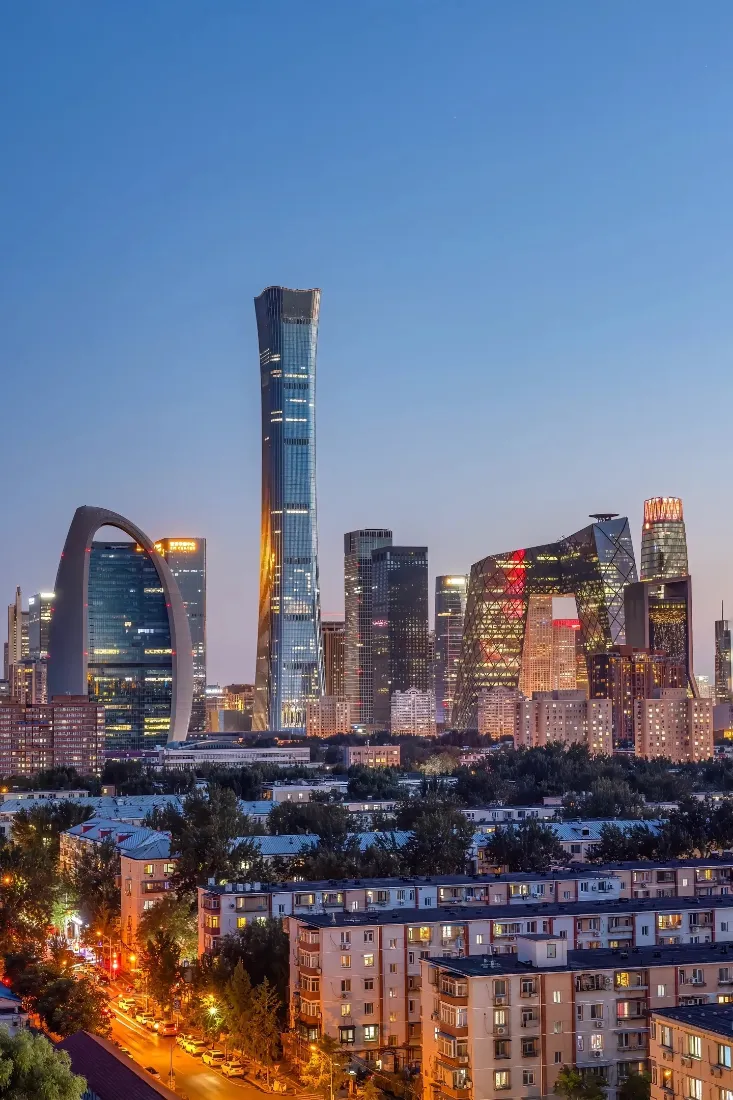 黄昏时分北京CBD天际线，中国尊矗立，建筑群灯火点亮，前景为居民区与街道。
