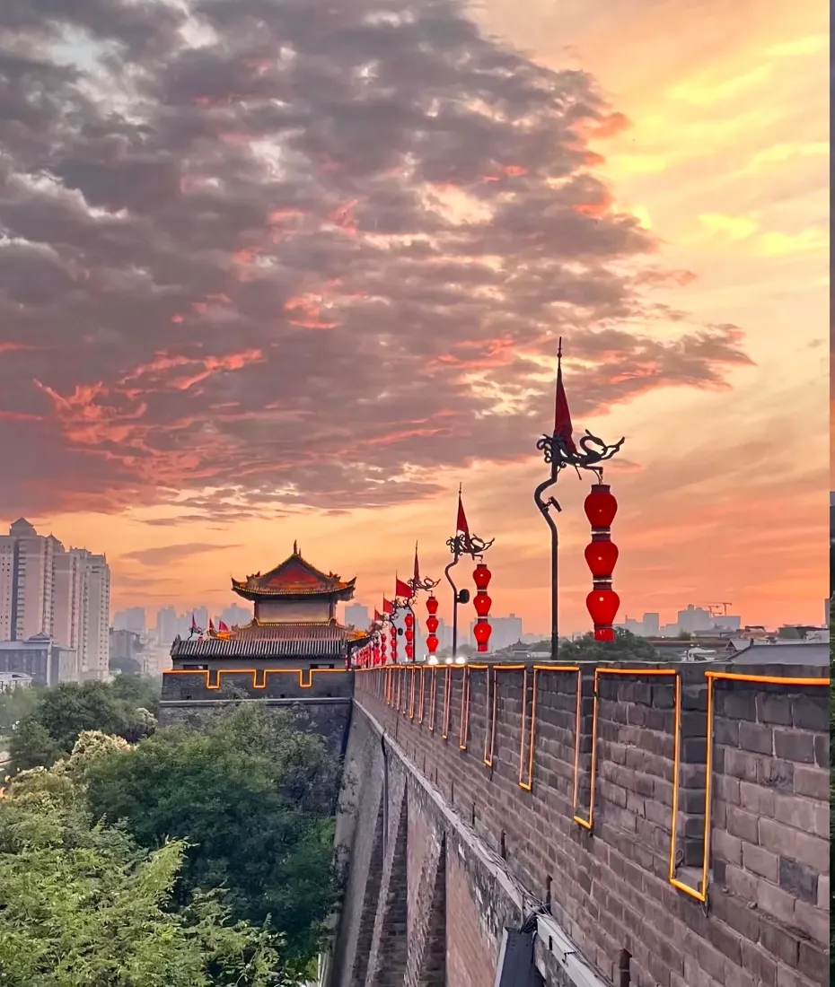 黄昏晚霞下的西安古城墙，沿墙悬挂红灯笼，远处可见城楼与现代城市建筑。