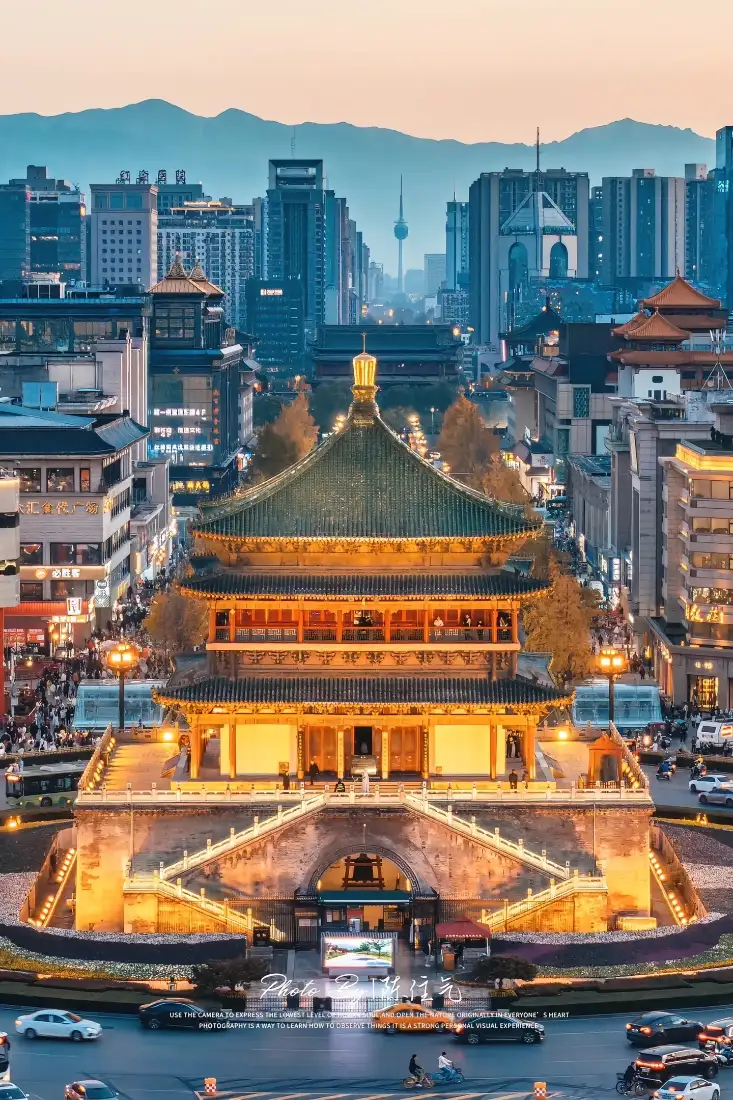 黄昏时分西安钟楼被暖黄灯光点亮，周边环绕现代都市建筑，远处可见连绵山脉。