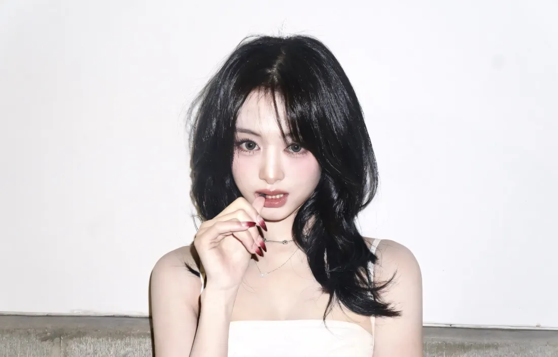 黑长卷发女子身着白色吊带，手抵唇边，背景为素白墙面