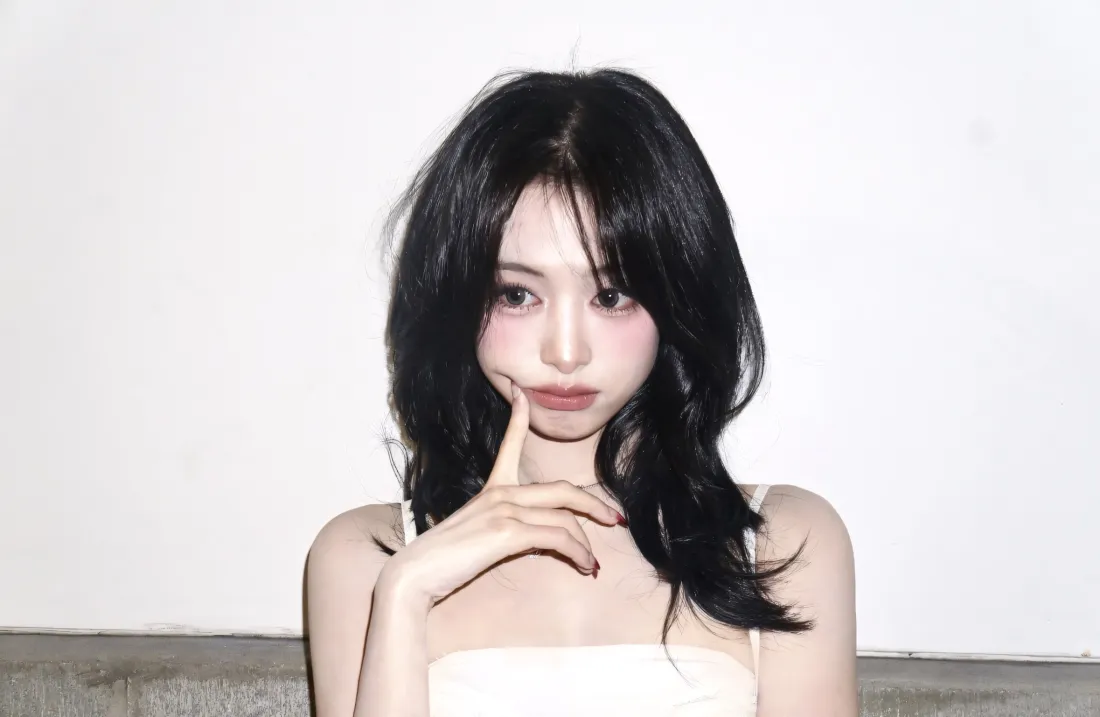 黑长发穿白色吊带的年轻女性，以手指轻触脸颊，背景为纯白墙面