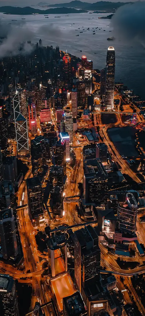 高空俯瞰的滨海都市夜景，楼宇与道路灯光交织，远处海面云雾缭绕。