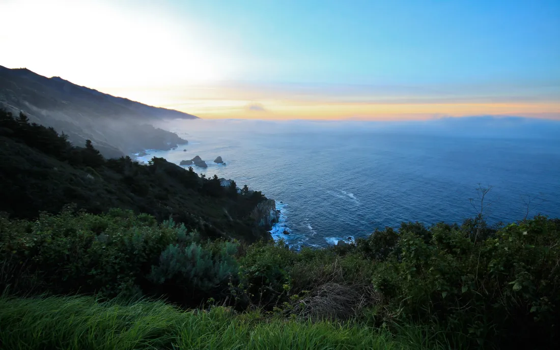 日出时分的海岸山景，俯瞰蓝色海洋与沿岸山峦，前景是茂密绿植