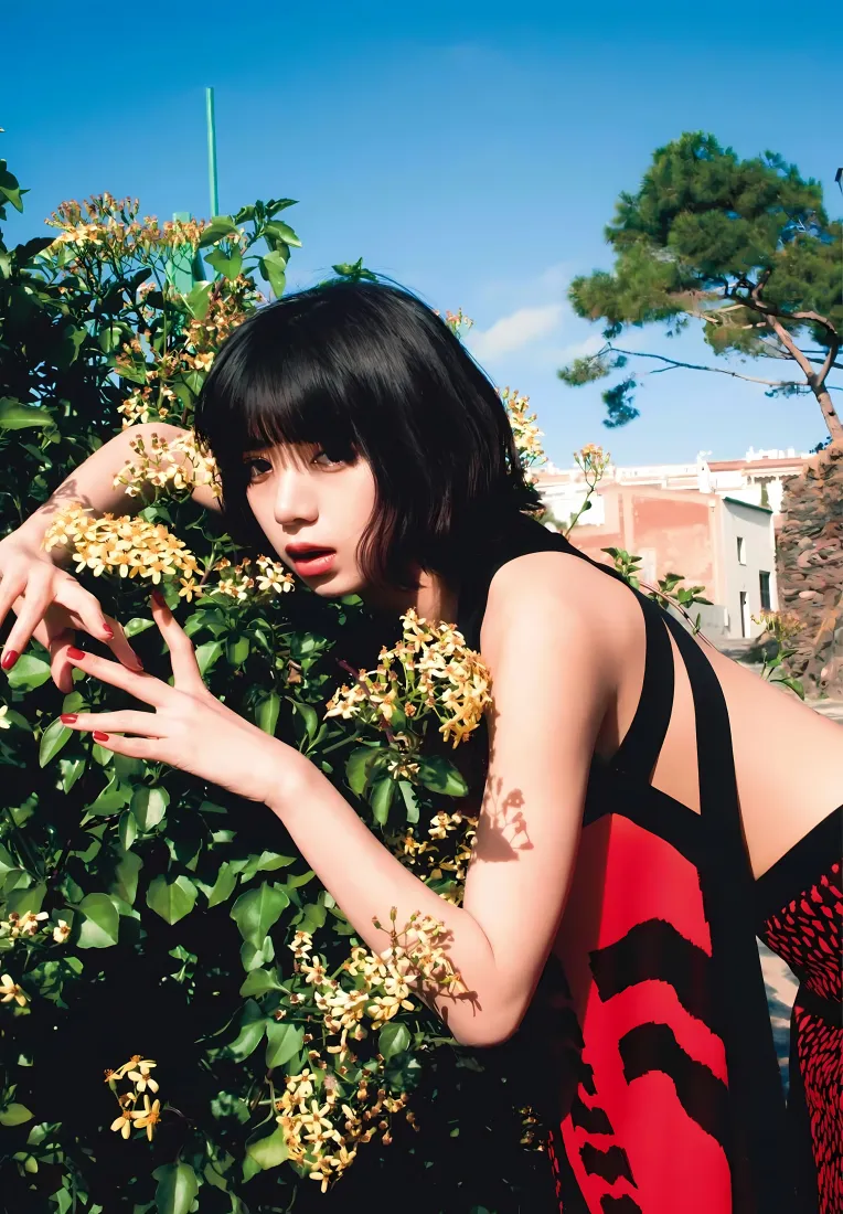 池田依来沙身着红黑吊带裙，倚靠在开黄花的绿植旁，背景是蓝天与远处建筑。