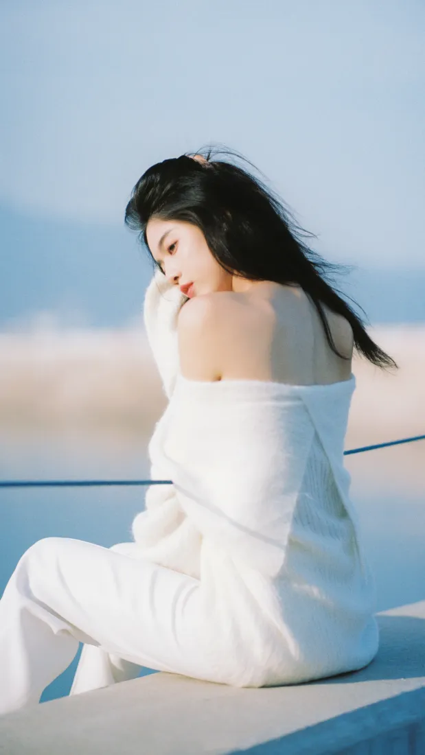 白衣露背长发女子坐于水边平台，背景为蓝天远山与水面。