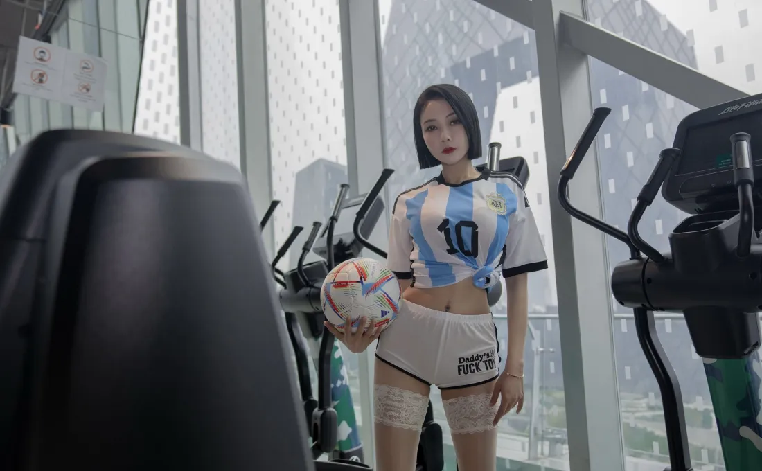 短发女子身着蓝白10号足球服，在健身房手持足球站立，背景为落地窗与健身器材。