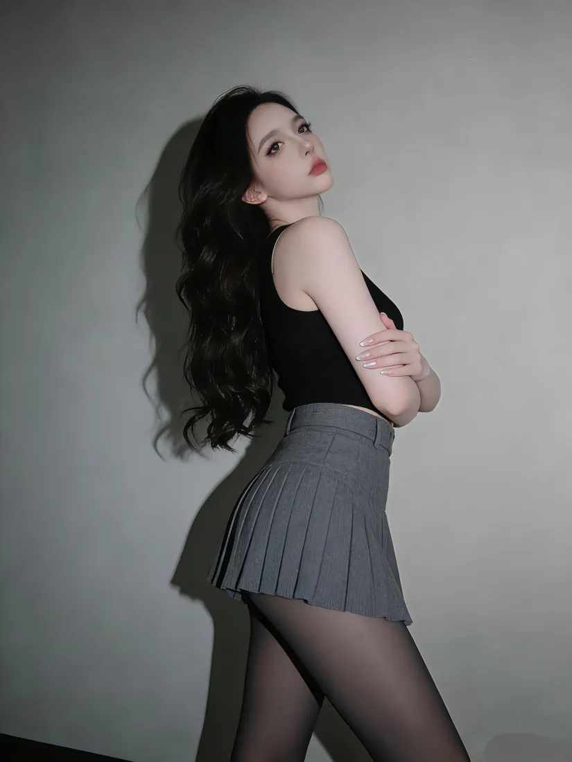 长发黑丝美女侧身倚靠浅灰墙面，穿黑色背心与灰色百褶短裙，双臂交叉。