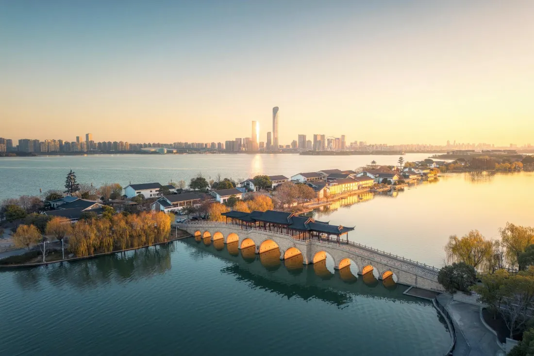 秋日日出时分，九孔廊桥横跨湖面，沿岸秋林与古建相映，远处城市天际线朦胧。