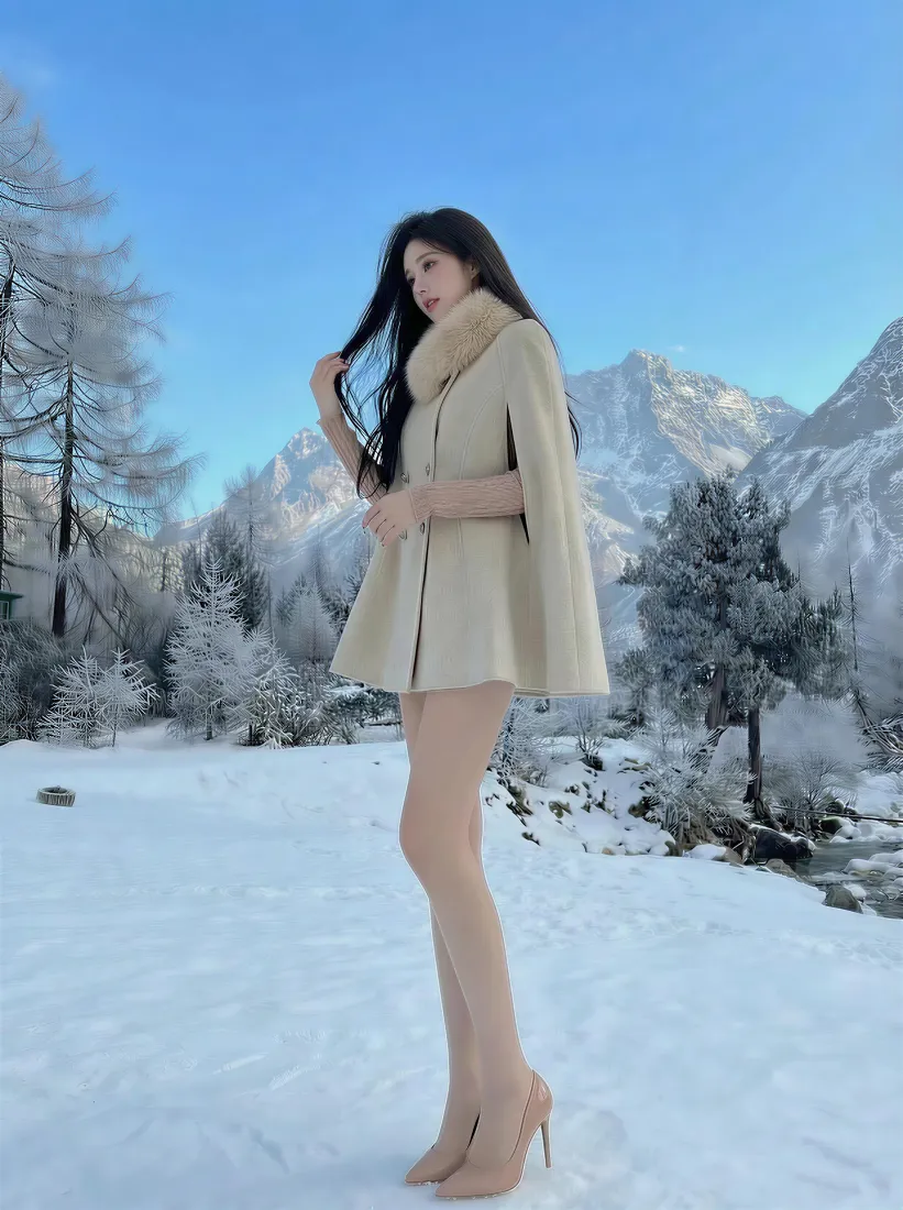 长发女子身着米色斗篷外套与高跟鞋，站在雪山前的雪地中。