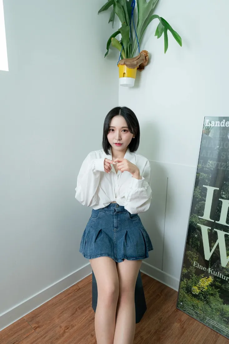 短发女生穿白衬衫蓝短裙坐于白墙木地板的居家角落，旁有绿植与海报。