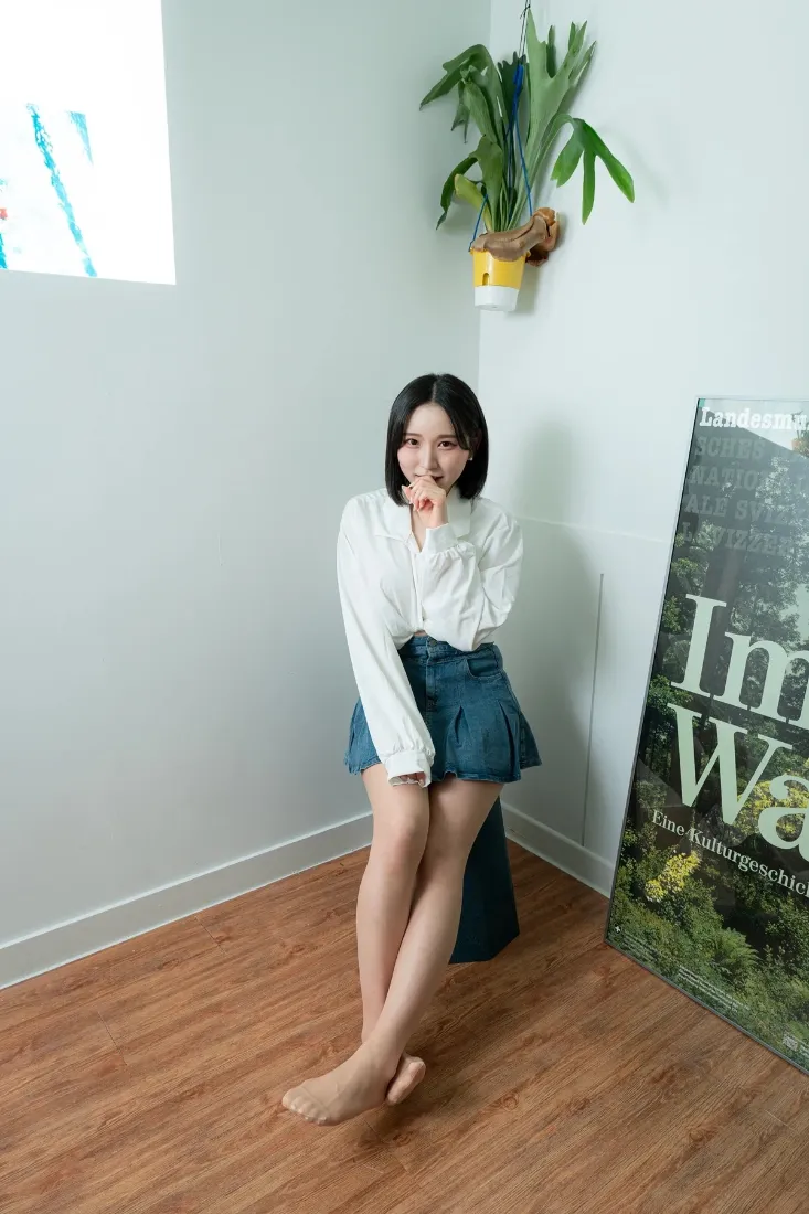 短发女生穿白衬衫蓝短裙光脚坐于白墙木地板的居家角落，旁有绿植与装饰画