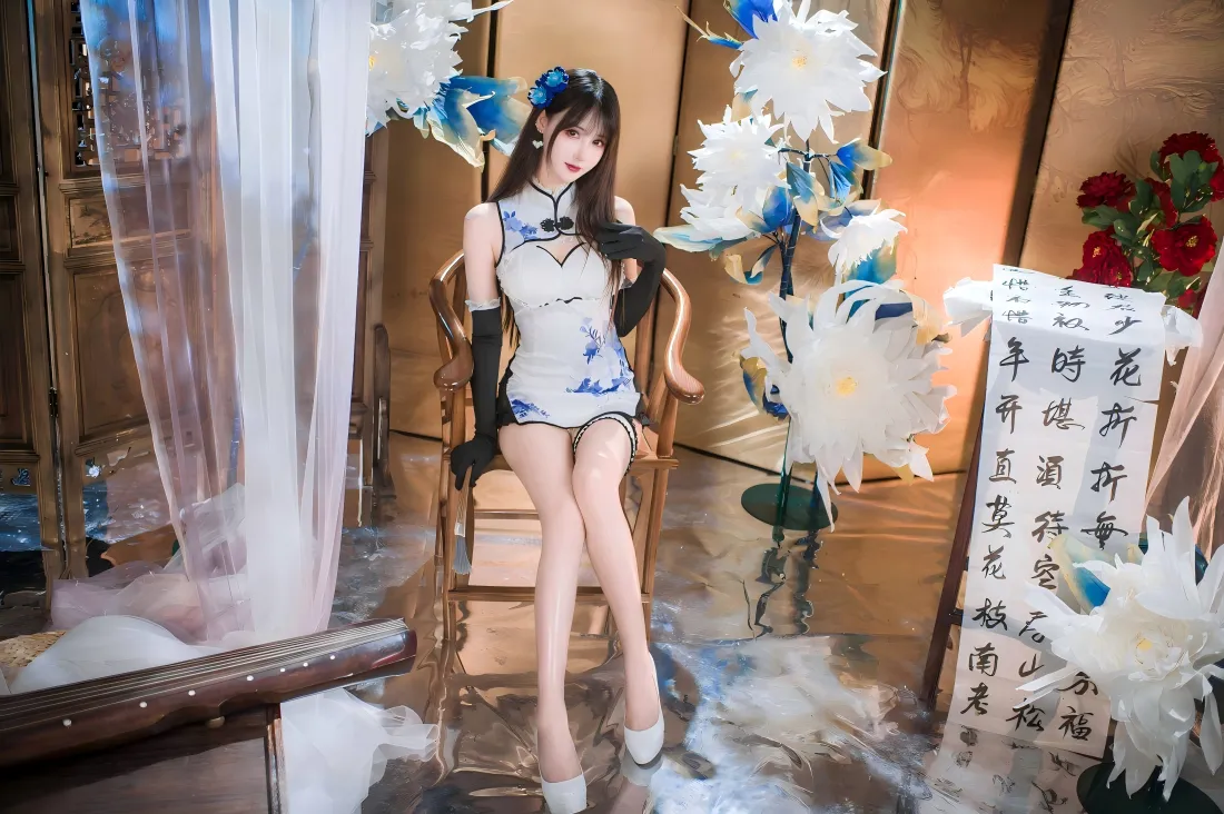 身着蓝白短款旗袍的女子坐于中式木椅，周围布置纸花与书法卷轴。