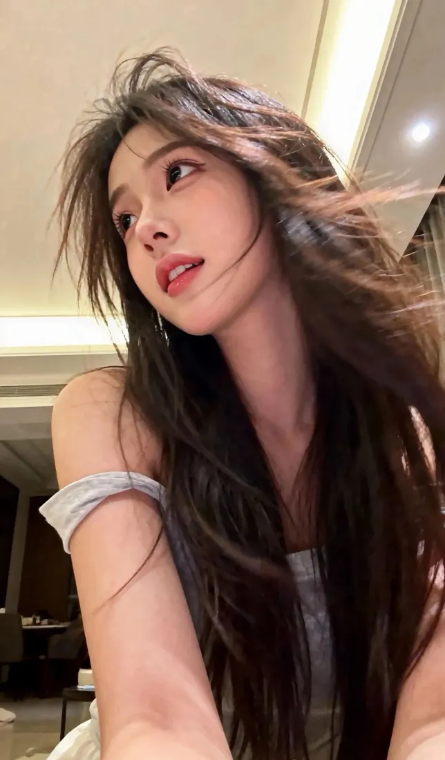 室内暖光下，长发年轻女子穿灰色露肩上衣，发丝飘动，抬眼望向上方。