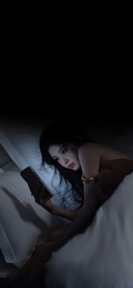 昏暗卧室里，年轻女子侧卧床上，手持手机，氛围静谧。