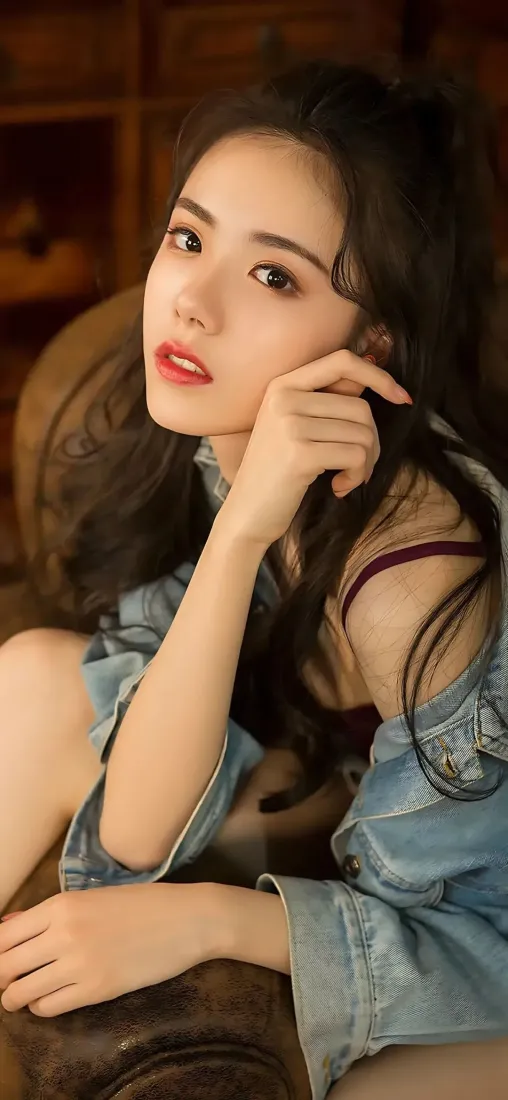 暖光室内，长发红唇女子穿露肩牛仔外套倚坐沙发，手托脸颊看向镜头。
