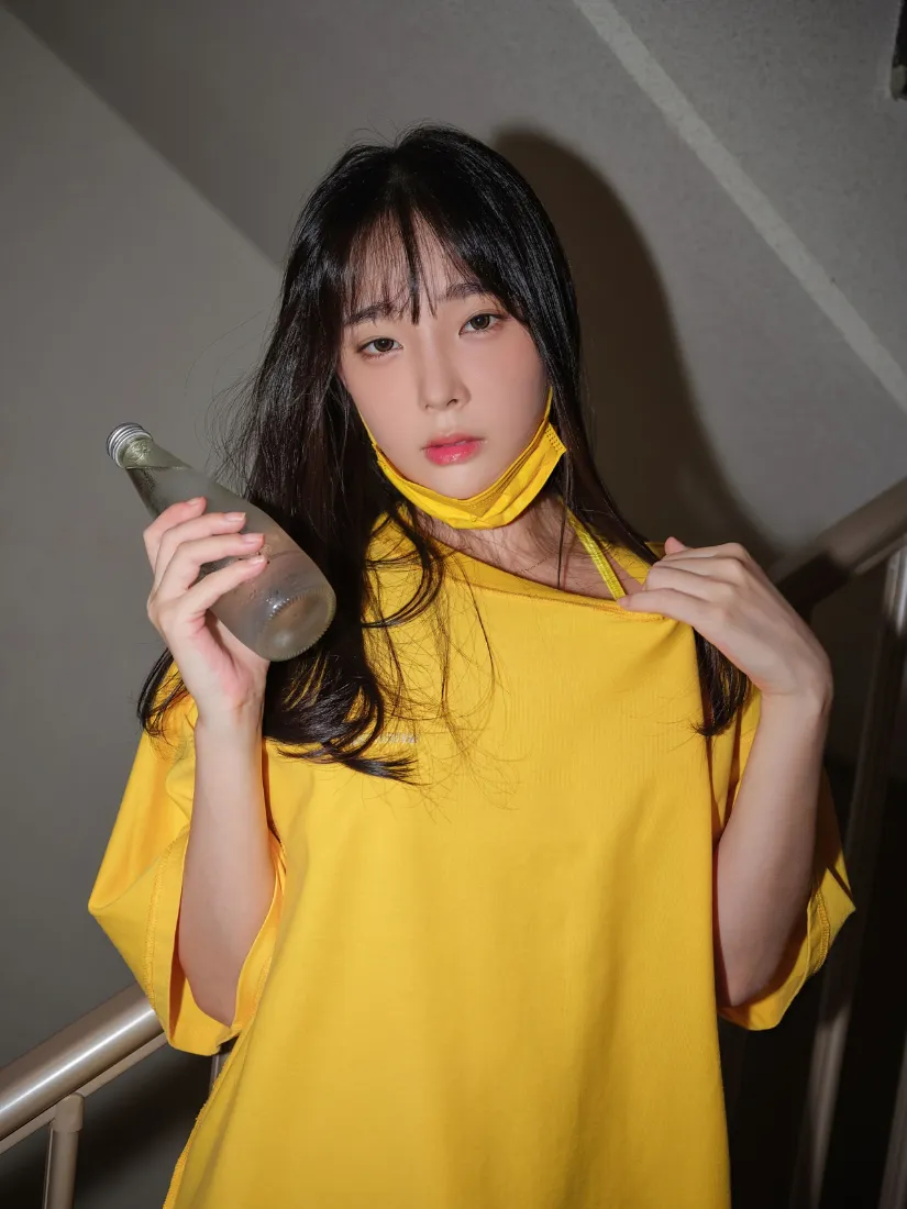 楼梯间内穿亮黄色上衣的长发女生，手持透明玻璃瓶，黄色口罩拉至下巴处。