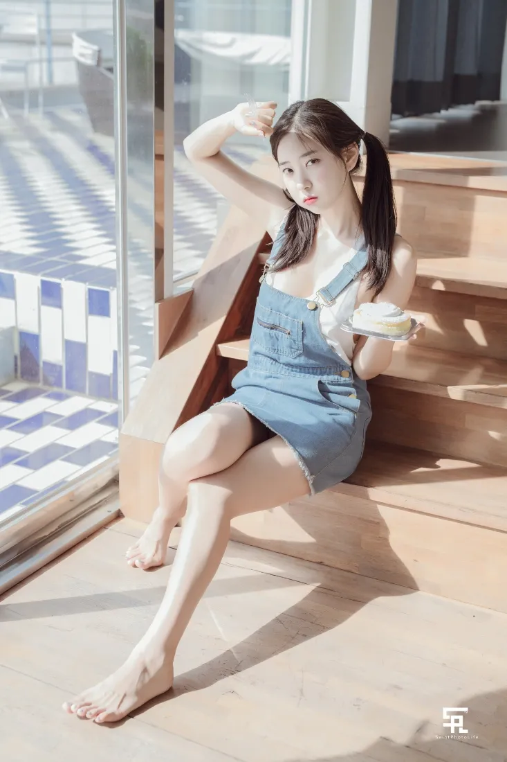 扎双马尾的少女穿牛仔背带裙，赤脚坐在阳光洒落的木质楼梯上。