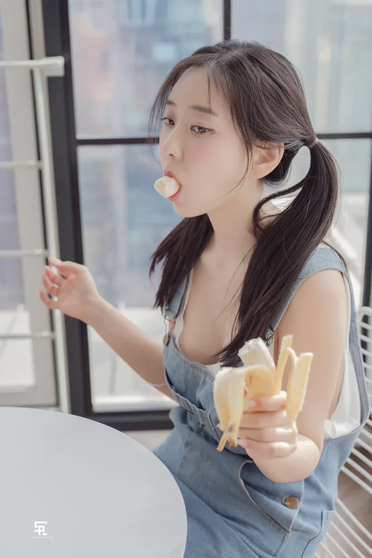 室内窗边，双马尾少女穿牛仔背带裙，手持香蕉正在进食。