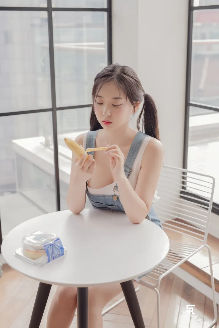 双马尾少女坐在明亮室内圆桌旁，手持香蕉，桌上摆放牛奶与蛋糕。