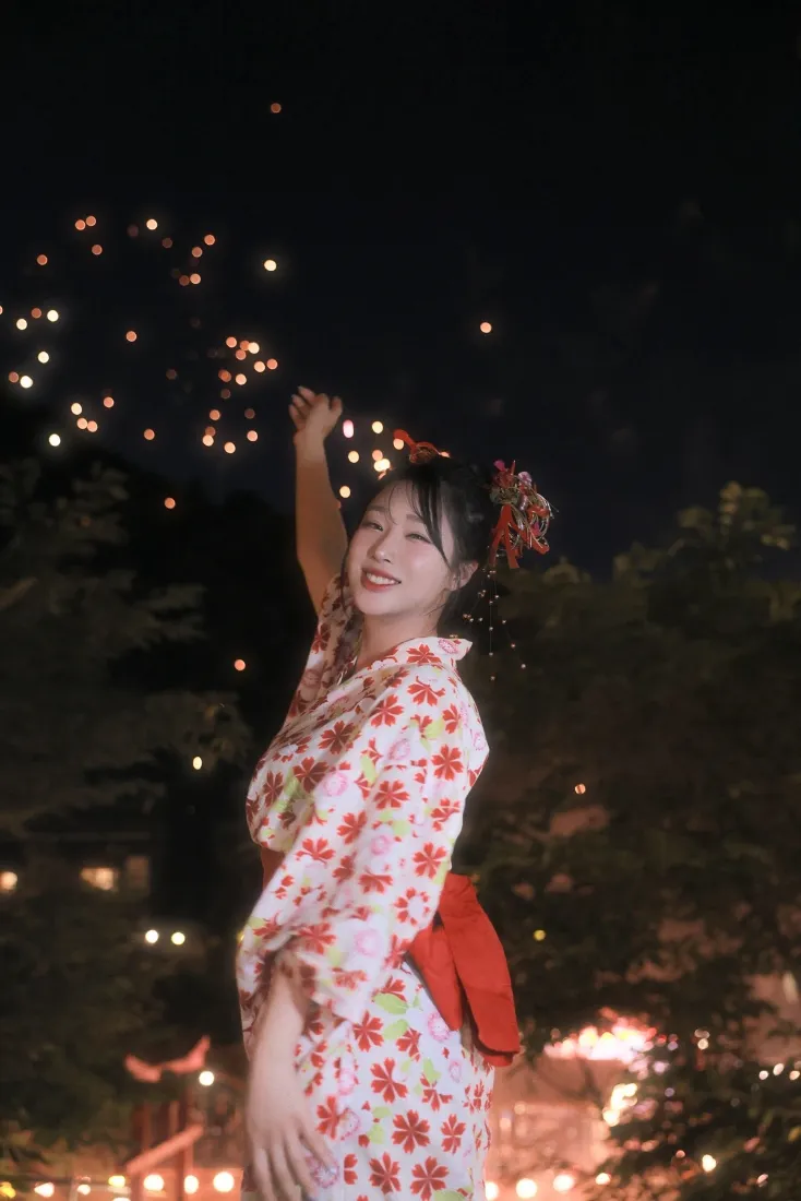 夜晚户外，身着红花和服配红腰带的少女抬手微笑，背景是虚化的暖光灯笼与树木。