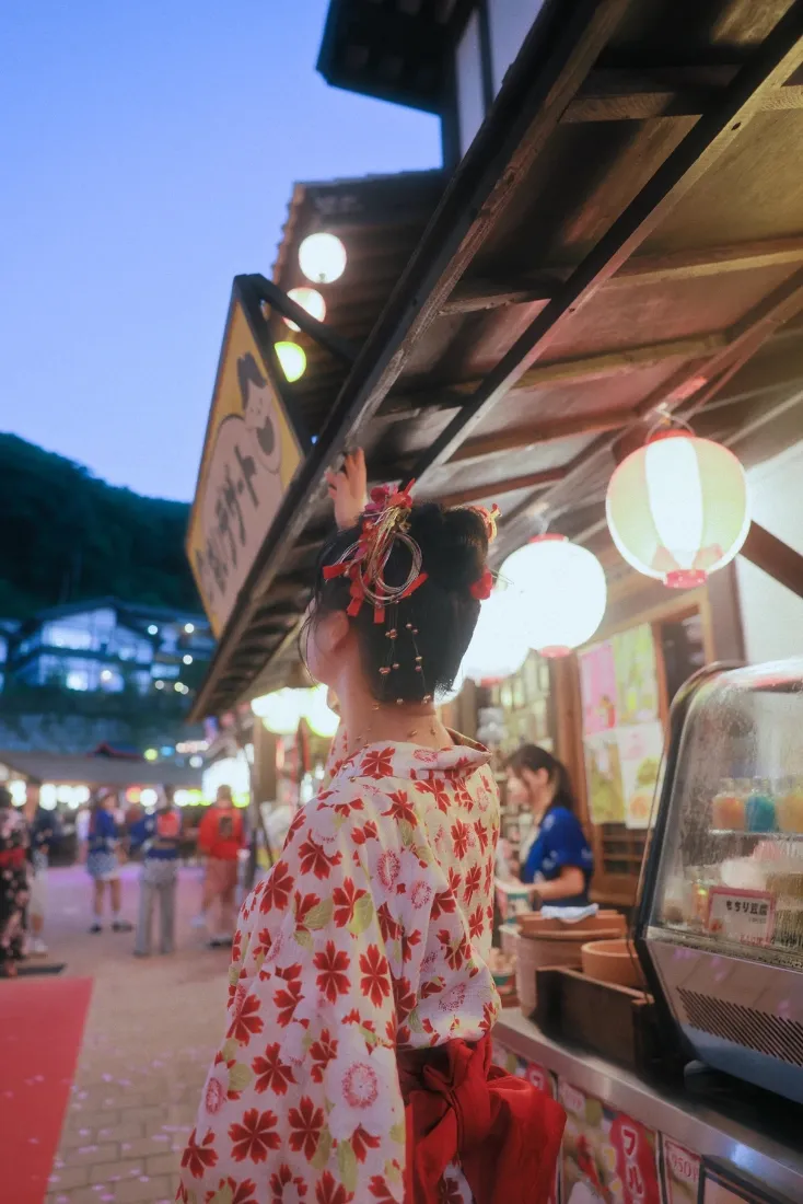 穿红色花纹和服的少女站在日式夜市摊位旁，黄昏蓝天下灯笼暖光柔和。