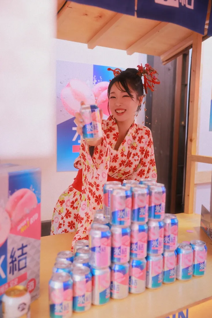 穿红色花纹和服的女子在饮料堆前举罐装饮料，笑容灿烂