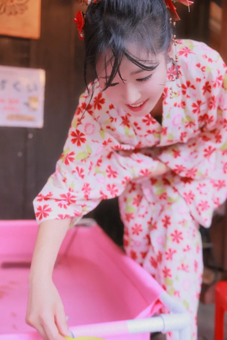 穿粉色红花和服的少女低头俯身，伸手触碰粉色容器，面带甜笑。