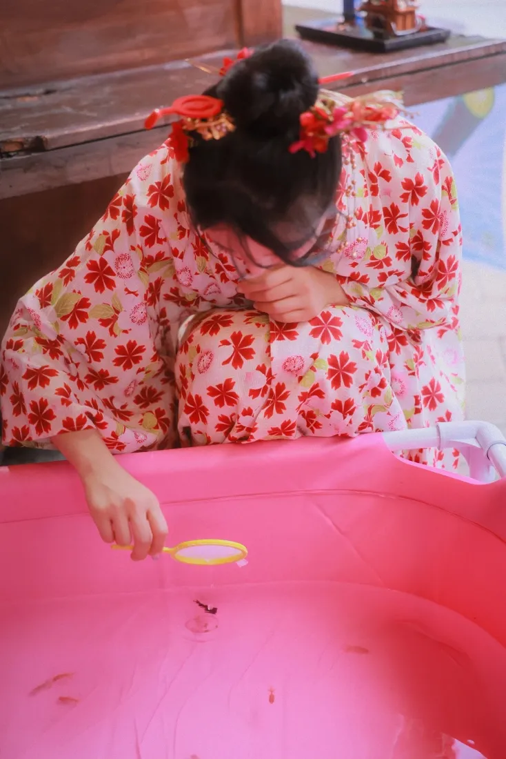 穿红白花纹和服的女子低头跪坐，手搭在粉色充气水池边。