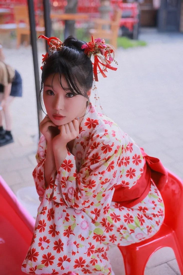 身着红色花纹和服的年轻女子坐于户外红色座椅上，双手托腮看向镜头。