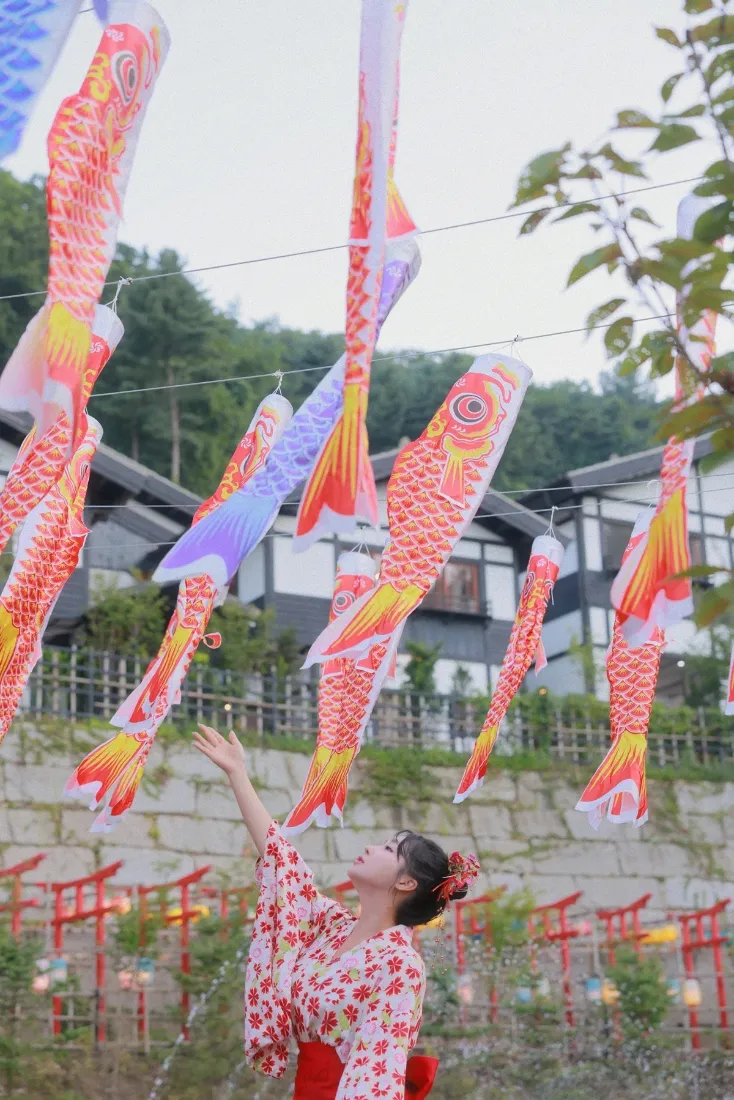 身着红纹和服的女子抬手仰望空中悬挂的多彩鲤鱼旗，背景为日式建筑与山林。