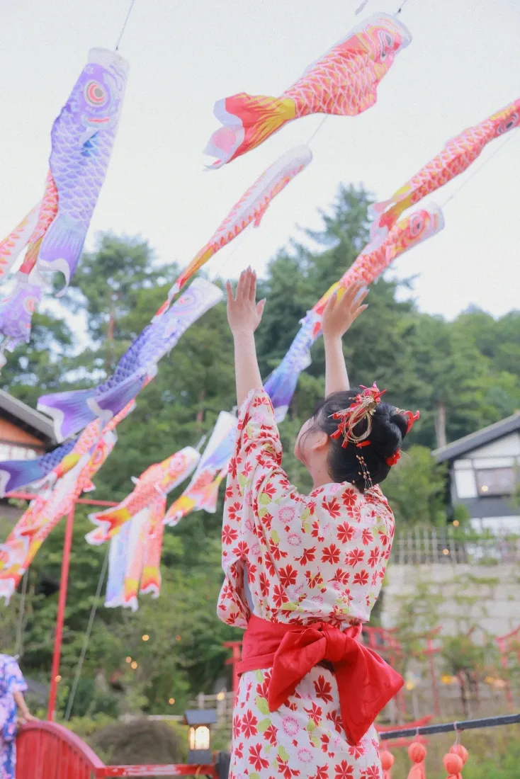 穿红樱花和服的少女背对镜头抬手，伸手触碰空中的彩色鲤鱼旗，背景是绿树与日式建筑。