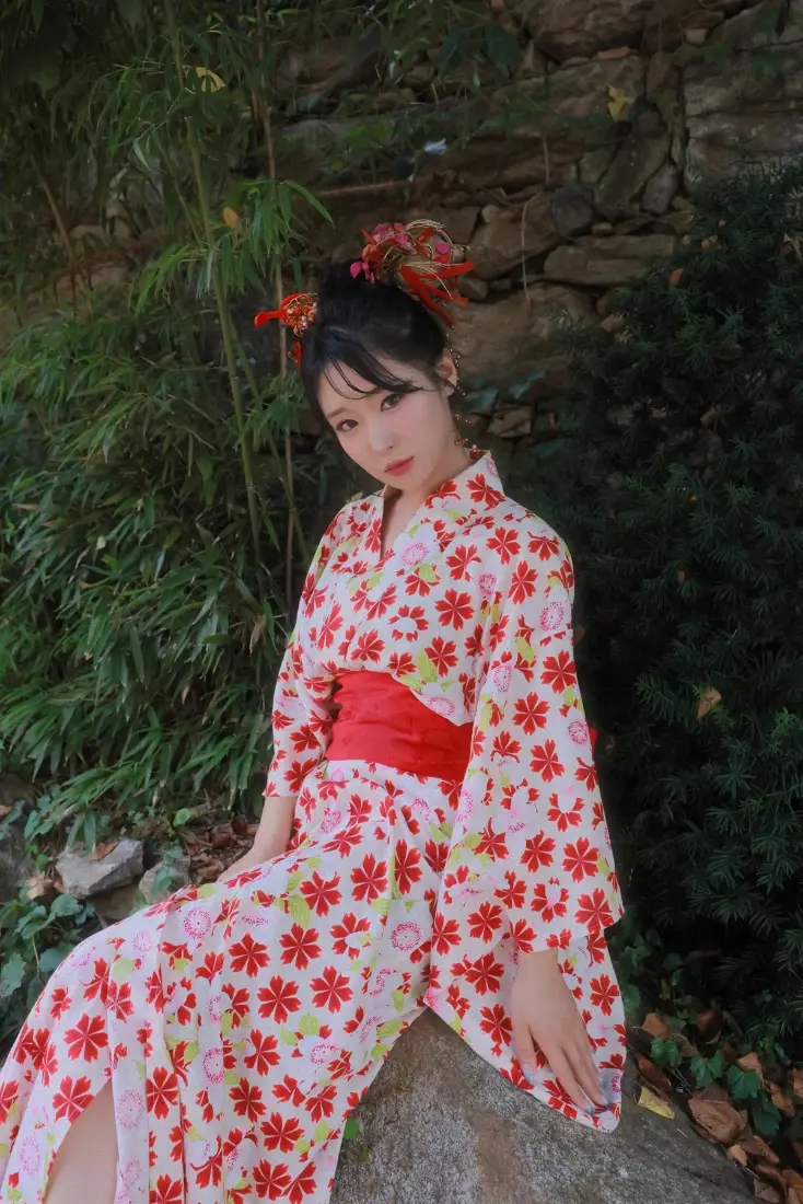身着红白花纹和服的少女坐在石上，背景为绿植与石墙。